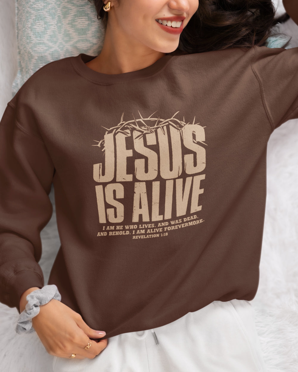a woman laying down in bed smiling, wearing a chestnut brown crewneck sweater with the words Jesus is Alive in a light brown color lettering with a crown of thorns on the top of the words Jesus.
