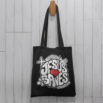 Black canvas tote bag with 'Jesus Saves' design hanging on a wooden hook against a light wooden panel background.