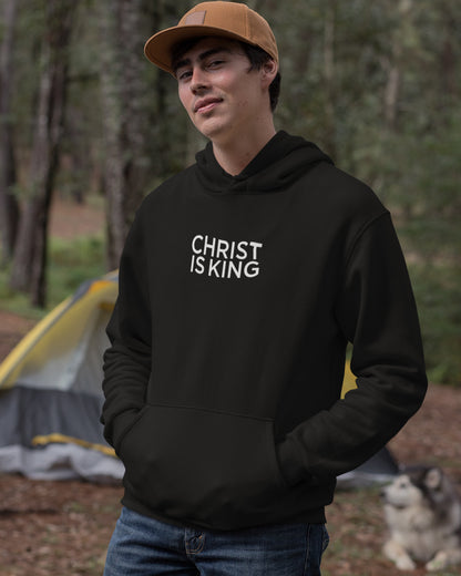 young man camping with a tent in the background in the forrest wearing a black hoodie that says Christ is King.