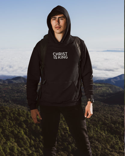 Person wearing a black hoodie with 'Christ is King' text, standing on a mountain with a scenic background.